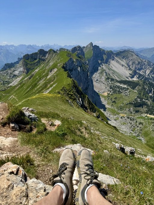 First-person view of hiking boots dangling over a rocky cliff edge, with a panoramic view of green alpine mountain peaks and valleys under a blue sky.