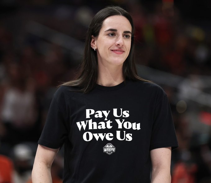A woman with long dark hair smiles while wearing a black t-shirt with white text reading 'Pay Us What You Owe Us' along with a WNBA logo.