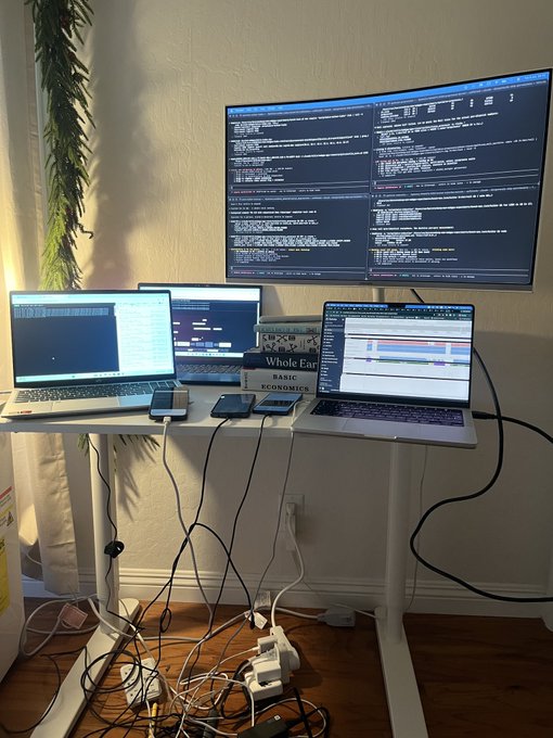 The image shows a desk cluttered with various electronic devices such as laptops, monitors, and cell phones. The desk has two large monitors displaying code, one of which is a web GPU. There are also multiple keyboards and mice scattered across the desk. A potted plant can be seen in the background, adding some greenery to the scene. The image seems to be related to a post about a person being locked into their computer for an extended period or a discussion on the challenges of working with electronic devices.