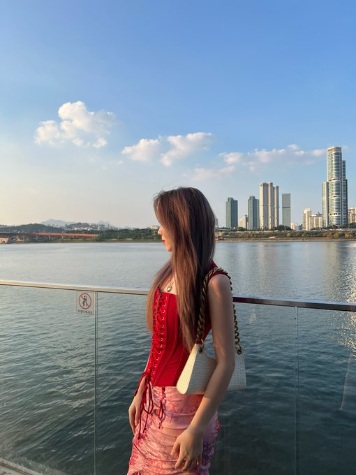 A woman in a red top and pink skirt stands by a waterfront railing, with a city skyline of tall buildings visible across the water.