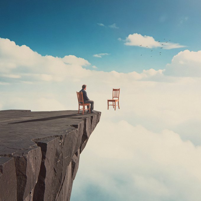 A man in a suit sits on a wooden chair at the edge of a rocky cliff, facing an empty chair across an endless sky of clouds, with a small flock of birds flying in the distance. The surreal scene visualizes the artist's description of "Inner Dialogue"—two chairs representing opposing sides of an internal argument, with the solitary figure embodying both perspectives in a silent, endless conversation suspended above the clouds.