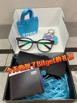 The image features a small box containing various items such as glasses, a wallet, and a keychain. The box also contains a blue bag with a white handle and a pair of glasses. The text "GiGi Fortune Pig and Bitget jointly remind you: enable multi-factor authentication and bind Google Authenticator Set up anti-phishing code: All official Bitget emails received will include your exclusive anti-phishing code For all types of assets, verify thoroughly". This message is related to the items in the box as it encourages users to take advantage of the provided security measures for their email account.