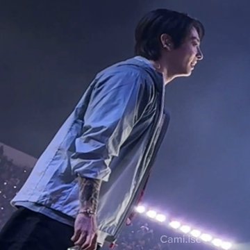 A male performer in a light blue jacket leans forward on a concert stage under blue lighting, with bright spotlights behind him and a faint “CamiLoc” watermark.