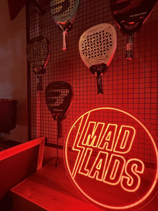 A red-lit interior corner featuring paddle rackets mounted on a wire grid wall next to a glowing red neon sign that reads "MAD LADS."