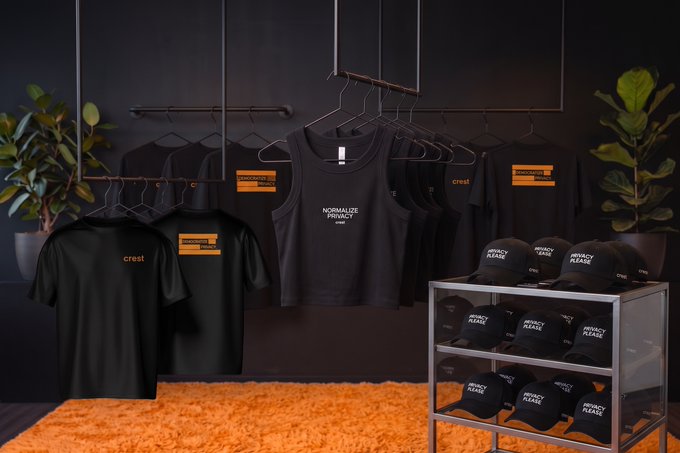 A merchandise display featuring black t-shirts and tank tops on hangers alongside stacked 'Cresti' branded caps, with apparel displaying 'NORMALIZE PRIVACY' and orange accents, set in a dark room with green plants and an orange rug.