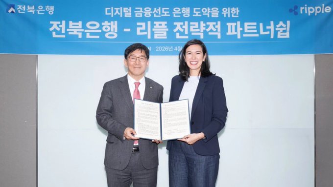 Two business professionals, a man and a woman, stand smiling and holding signed documents in front of a blue backdrop featuring Korean text and the Ripple logo, indicating a strategic partnership.