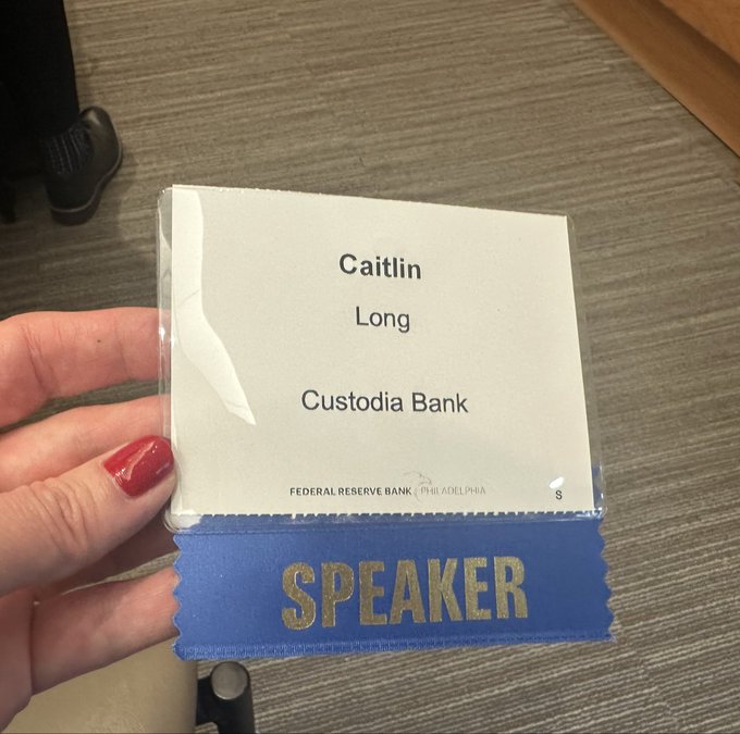 A hand holds a speaker badge for Caitlin Long of Custodia Bank at the Federal Reserve Bank of Philadelphia, ready for her presentation as noted in the caption highlighting the packed in-person room and 1,700 online registrants.