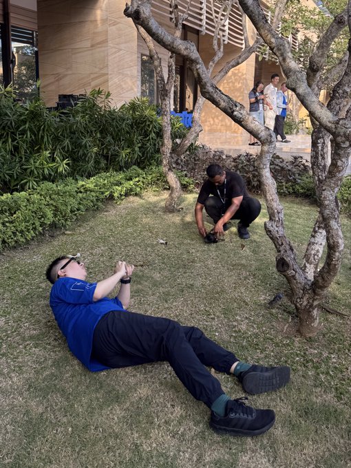 Two men in an outdoor courtyard area; one lies on the grass holding a phone while the other crouches nearby examining something on the ground.