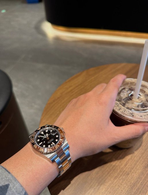 A person's wrist wearing a Rolex GMT-Master II watch with a black and brown bezel, next to a hand holding an iced coffee drink on a wooden table.