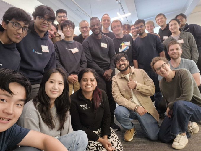 A group photo of approximately 15-20 diverse people, with one person wearing a sweatshirt with the 'bryanjb' logo, posing together indoors.