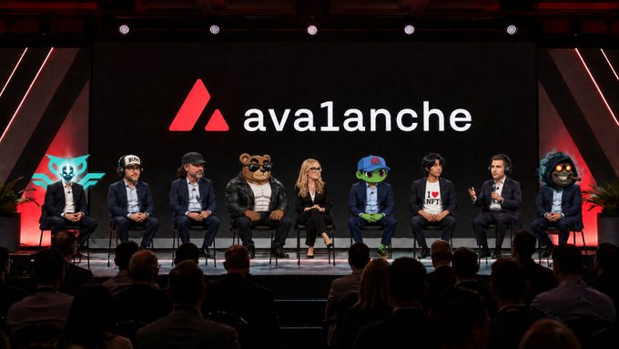 The image features a group of people sitting around a stage with a large screen behind them displaying the word "avalanche" written in white letters. The audience consists of several individuals, including some wearing suits and ties, who appear to be engaged in a discussion or presentation. One person is holding up a teddy bear while another person has a green frog on their lapel. The stage setting suggests that this gathering could be related to the cryptocurrency industry or a similar field where Avalanche's impact is being highlighted.