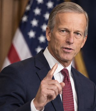 A man in a dark suit and red patterned tie gestures with his index finger raised, speaking in front of an American flag.