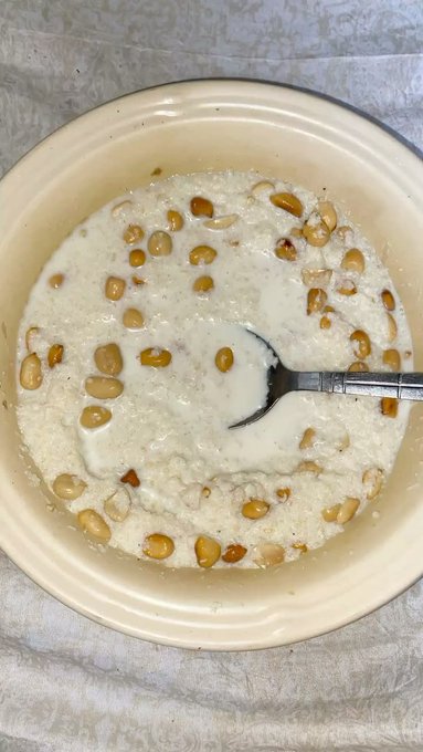 A cream-colored ceramic bowl filled with a creamy dish topped with soybeans or nuts, with a metal spoon resting inside, placed on a white patterned cloth.