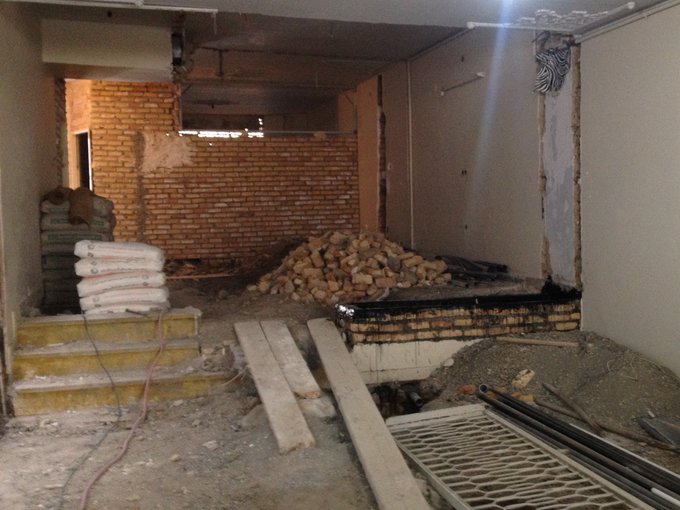 An unfinished basement under renovation with exposed brick walls, stacked bags of construction material, a pile of rubble, and wooden planks scattered on the dirt floor.
