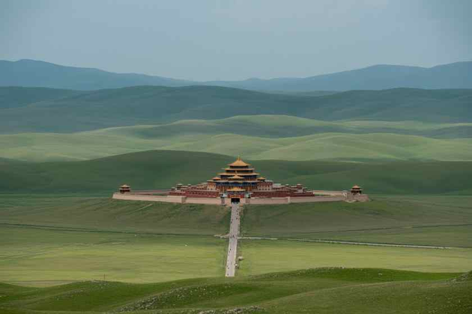 A large traditional Asian-style temple complex with golden roofs sits on a grassy plain, surrounded by rolling green hills and mountains, with a long pathway leading up to it.