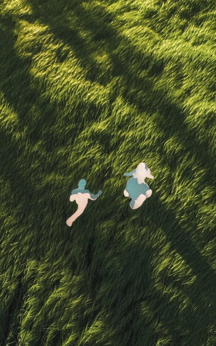 The image features two people standing in a field of tall grass, both wearing blue shirts. The person on the left has their back turned towards the camera while the other person is facing forward. They appear to be enjoying themselves as they stand together in the green grassy area.