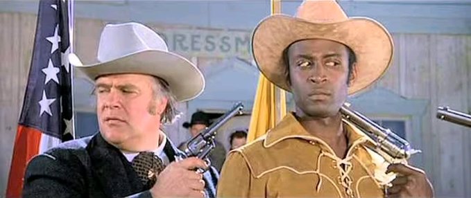 Two cowboys in a standoff—one older white man in a gray hat pointing a revolver, and a younger Black man in a tan hat pointing his own pistol back—evoke the tense "let's not negotiate with thieves" dynamic of the Bitcoin bug standoff described in the post. The "RESS M" sign and American flag in the background reinforce the Wild West "fix the bug or else" metaphor.