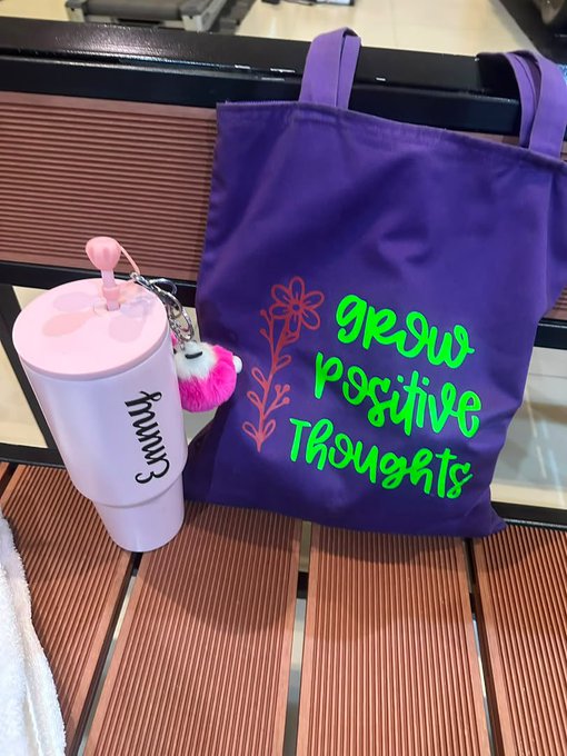 A pink tumbler with the name 'Emma' and a purple tote bag displaying 'Grow Positive Thoughts' in green text with floral designs, sitting on a wooden bench.