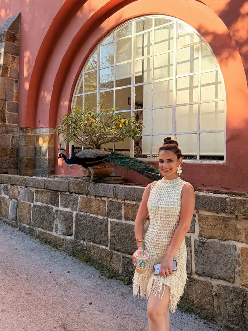 A young woman in a cream-colored fringed dress smiling while holding a drink and phone, posing in front of a large arched window with a peacock perched on a planter beside her.