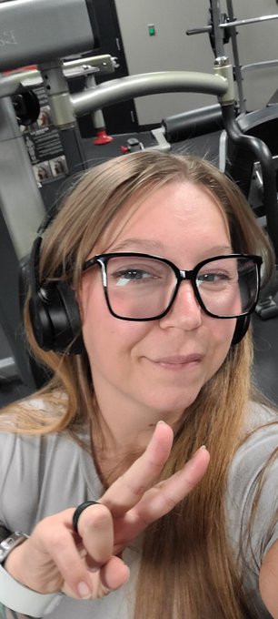 A selfie of a woman with glasses and headphones in a gym, smiling and holding up a peace sign.