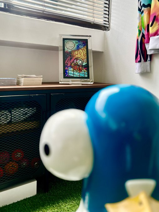 A blurred blue character statue in the foreground looking toward a framed 'BAD ART' digital artwork displayed on a dark cabinet in a room with artificial grass.