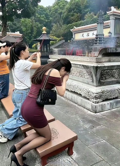 A woman in a maroon off-shoulder dress and black high heels kneels on a bench at a temple, bowing before an incense-filled urn, while another woman in the background takes a photo.