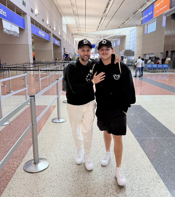 Two smiling men stand in an airport terminal, both wearing black hoodies with matching 'D' logo baseball caps, with one having his arm around the other.