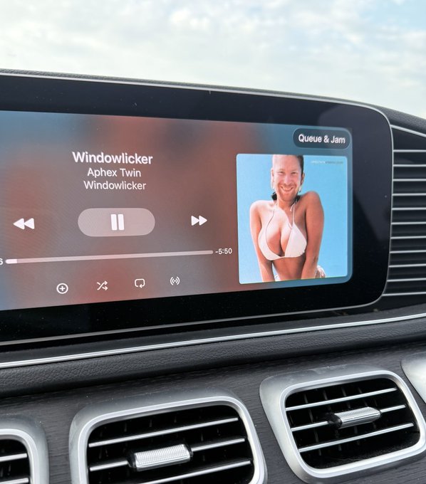 A car's infotainment screen displaying Apple Music, currently playing 'Windowlicker' by Aphex Twin on the 'Queue & Jam' section, with album art showing a smiling woman in a bikini.