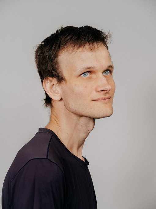 A portrait of a young man with brown hair and blue eyes, wearing a dark shirt, photographed against a light gray background.