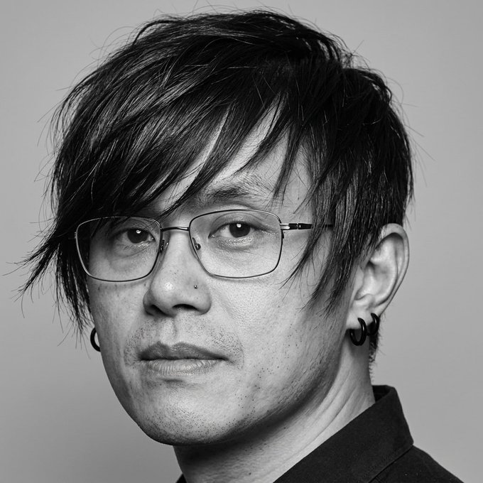 A black-and-white portrait photograph of an East Asian man with medium-length dark hair swept to one side, wearing rectangular wire-rimmed glasses, small hoop earrings in both ears, and a nose piercing, looking directly at the camera.