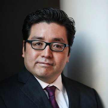 A professionally dressed man with dark hair and glasses, wearing a dark suit and tie, appears in a portrait-style photo. The image likely accompanies news about Tom Lee's BitMine making a significant Ethereum purchase of 27,180 ETH, as he is associated with Fundstrat and cryptocurrency investment analysis.