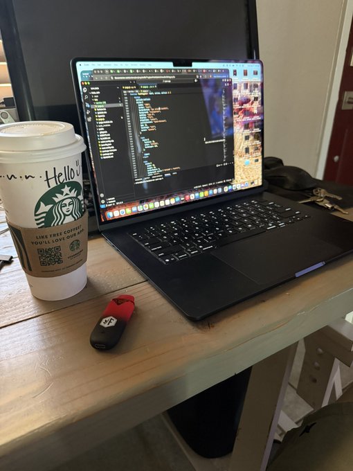 A laptop showing code on screen sits on a wooden desk next to a Starbucks coffee cup and a red USB drive.