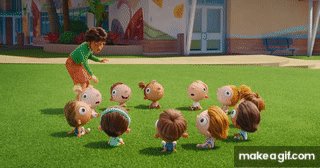 The image features a group of cartoon characters gathered together on a grassy field. There are at least 12 characters, including a girl standing near them with her arms crossed, and other children playing around her. The scene appears to be set outdoors during daytime hours. In the background, there is a building visible behind the group of characters.