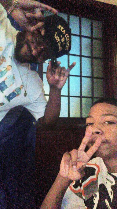 Vintage-style photo of two people, one wearing a 'BLAQ POET' cap making hand gestures, with another person in the foreground flashing a peace sign.
