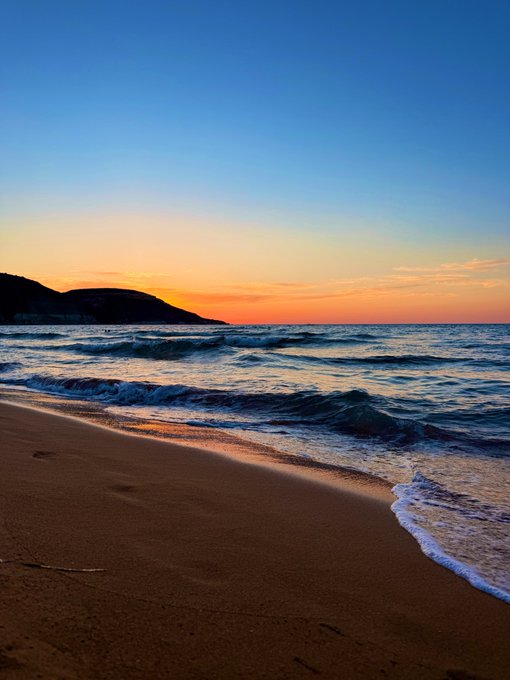 A serene beach at sunset, with waves gently rolling onto the sandy shore under a gradient sky of deep blue and warm orange hues, complemented by a distant silhouetted coastline.

The image pairs with the caption "gn bulls, I want you to make enough this cycle to buy more time with the people you love" by evoking the value of trading success—not for wealth itself, but to reclaim peaceful, meaningful moments like watching a sunset by the ocean with loved ones.