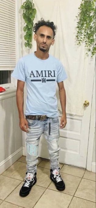A man stands indoors wearing a light blue 'AMIRI' t-shirt, distressed light-wash jeans with 'ER' and 'CA' lettering on the legs, and black and white high-top sneakers.