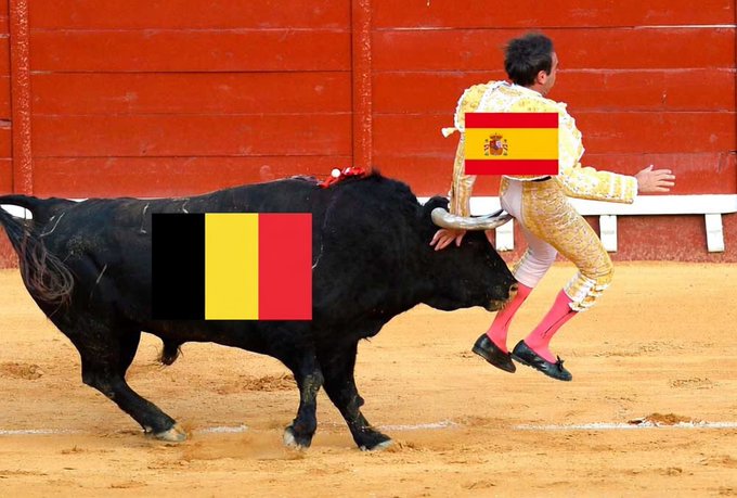 A bullfighting scene with a Spanish flag emoji placed over the bull and another Spanish flag emoji on the matador, while a small black-yellow-red Belgian flag is overlaid on the bull's body.