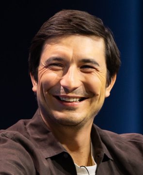 A close-up portrait of a smiling man with dark hair wearing a brown shirt against a dark blue background.