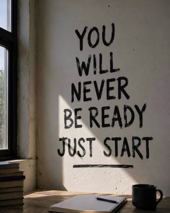Black hand-painted text on a white wall reads 'YOU WILL NEVER BE READY JUST START', next to a window with stacked books, a notebook, pen, and black mug on a desk.