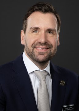 A professional headshot of a man in a dark suit, white shirt, and silver tie with a name badge reading 'JASON DUKES' against a gray background.