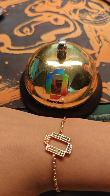A person wears a gold "renaiss" bracelet on their wrist, displayed next to a gold service bell with the Renaiss logo, celebrating receiving the chain as a gift from the Renaiss brand.
