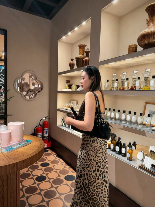 A woman in a leopard-print skirt stands in a perfume shop examining bottles on illuminated shelves, with a fire extinguisher visible nearby.
