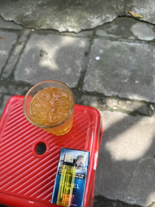 A glass of iced tea and a yellow vape device resting on a red plastic crate on a stone-paved ground in the sunlight.