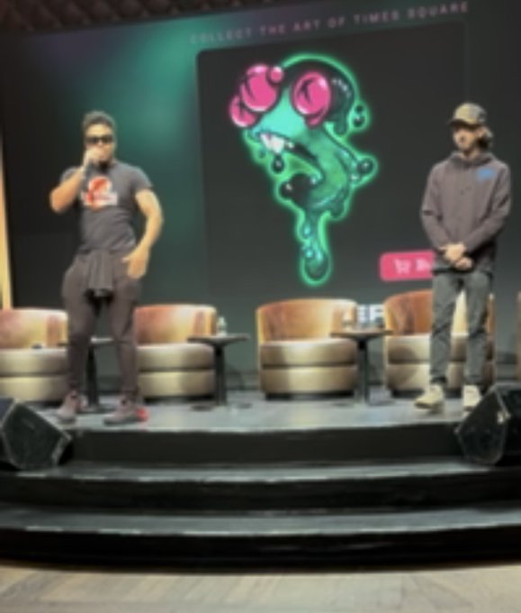 Two people stand on a stage at a Times Square art event, with one holding a microphone and a screen behind them displaying 'COLLECT THE ART OF TIMES SQUARE' alongside a colorful cartoon creature.