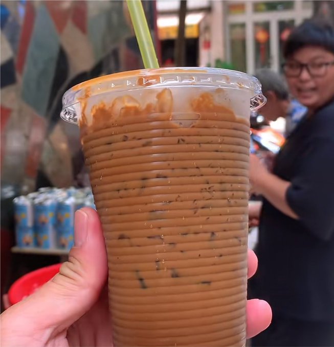 A hand holds a clear plastic cup of Thai iced milk tea with a green straw, showing the characteristic brown tea color with visible ice and the orange Thai tea staining the inner lid.