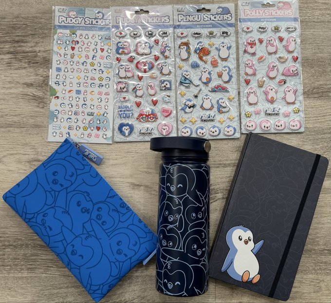 An assortment of penguin-themed merchandise is laid out on a wooden table, featuring four packs of puffy stickers, a blue patterned zip pouch, a black thermos with penguin line art, and a black notebook with a cute penguin design.
