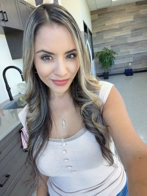 A selfie of a woman with long wavy brown and blonde highlighted hair, wearing a white sleeveless top and a small cross necklace, standing in what appears to be a kitchen or break room with wooden cabinets.