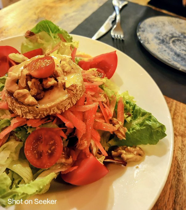 A photograph of a plated salad featuring mixed greens, cherry tomatoes, shredded carrots, walnuts, and a round cheese disc, with "Shot on Seeker" watermark at the bottom.
