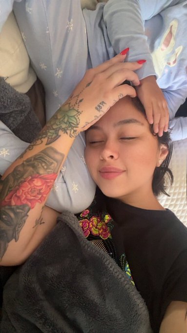 A young woman with closed eyes and a serene expression rests her head while another person's tattooed arm and hand (with red painted nails) rests on her forehead.