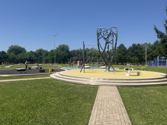 An outdoor park scene featuring a modern playground with a large silver metal tree-like sculpture, yellow safety surfacing, exercise equipment, and people enjoying the sunny day.